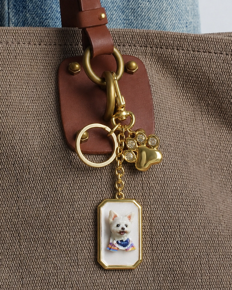 Gold keychain with a 3D white dog portrait and paw charm clipped to a brown canvas tote bag.