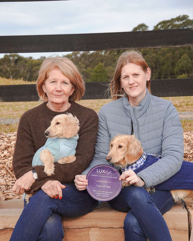 sarah and jane sitting outdoors holding small dachshunds in sweaters and a round purple award plaque.
