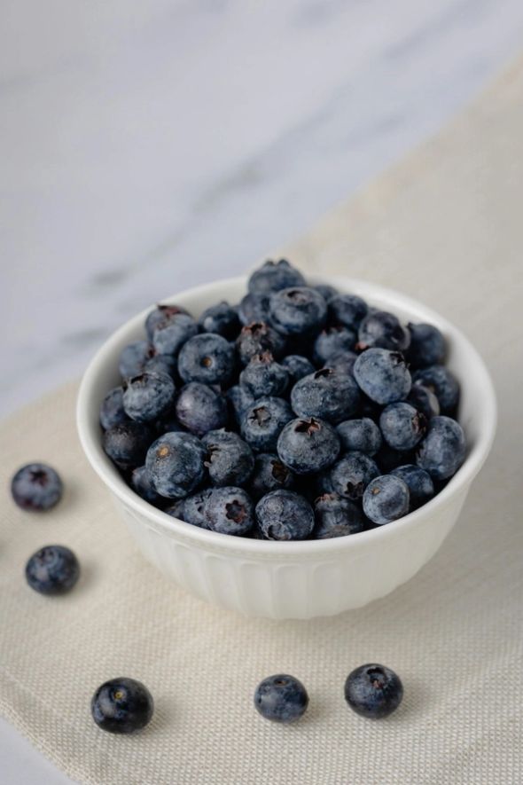 A white bowl filled with blueberries, with several loose berries scattered on a beige cloth.