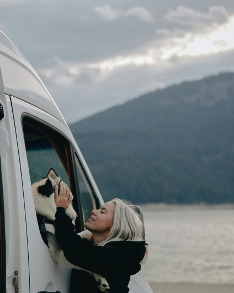 A dog mum petting her husky looking out of a camper van window while RV camping with her dog by a scenic lake.