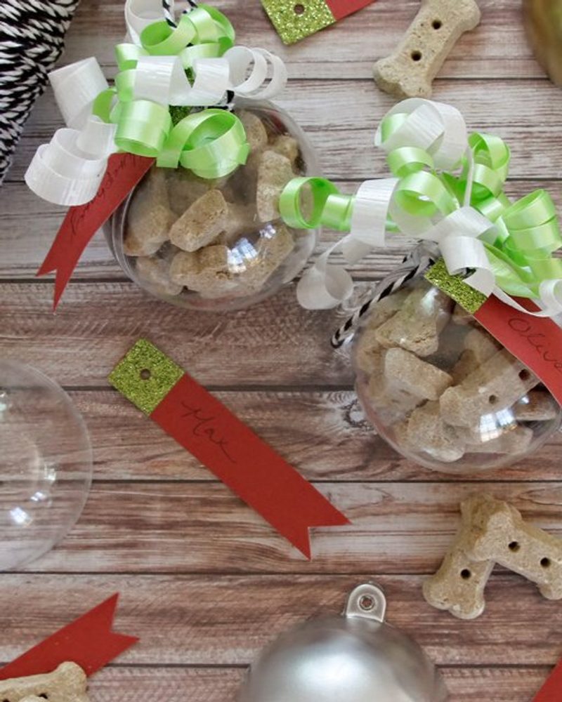 DIY dog treat bauble ornaments filled with bone-shaped biscuits, featuring green ribbons and personalized name tags.